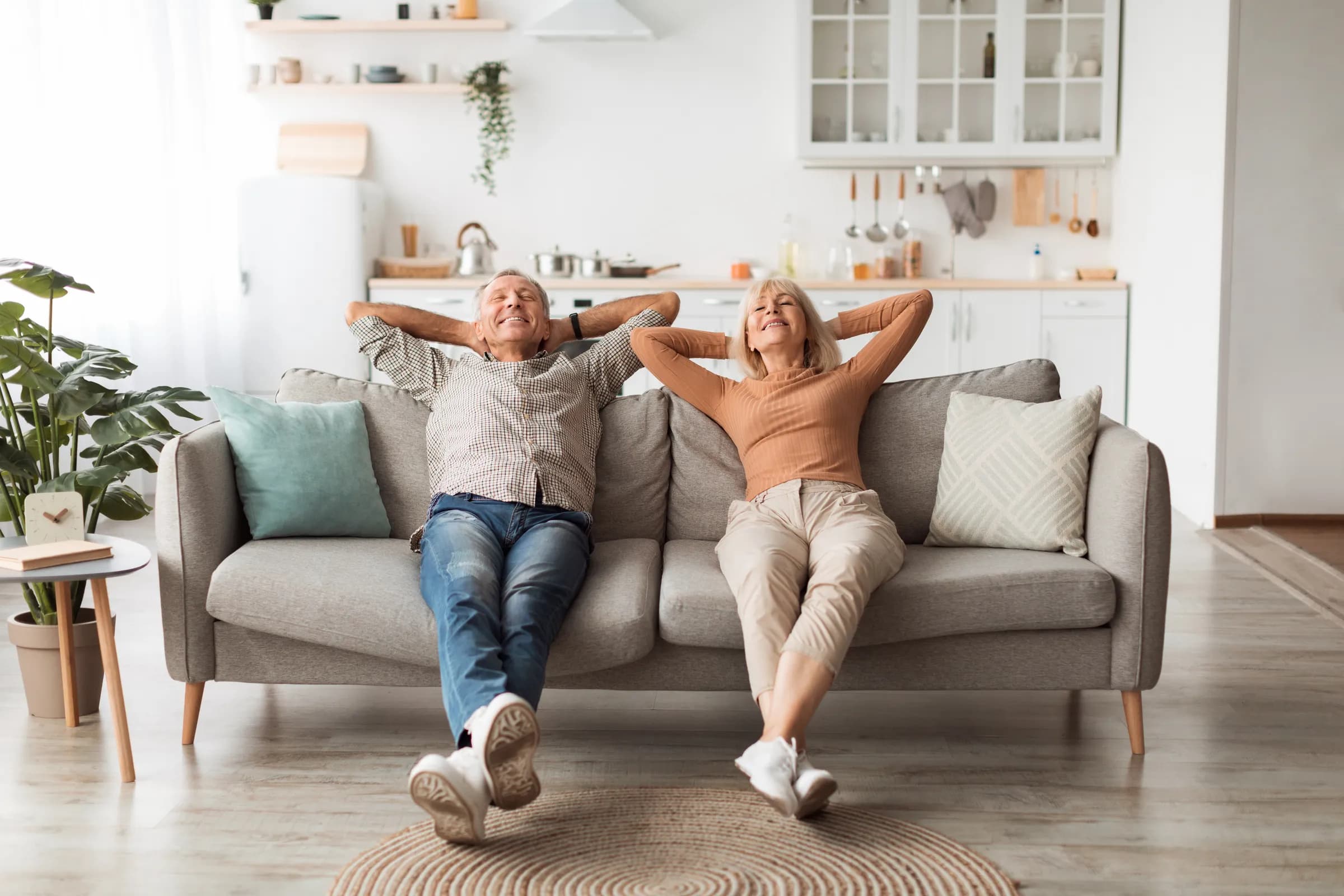 Couple de seniors détendu, mains derrière la tête, sur le canapé de leur salon clair — bien vivre chez soi avec les soins à domicile Vitaloria