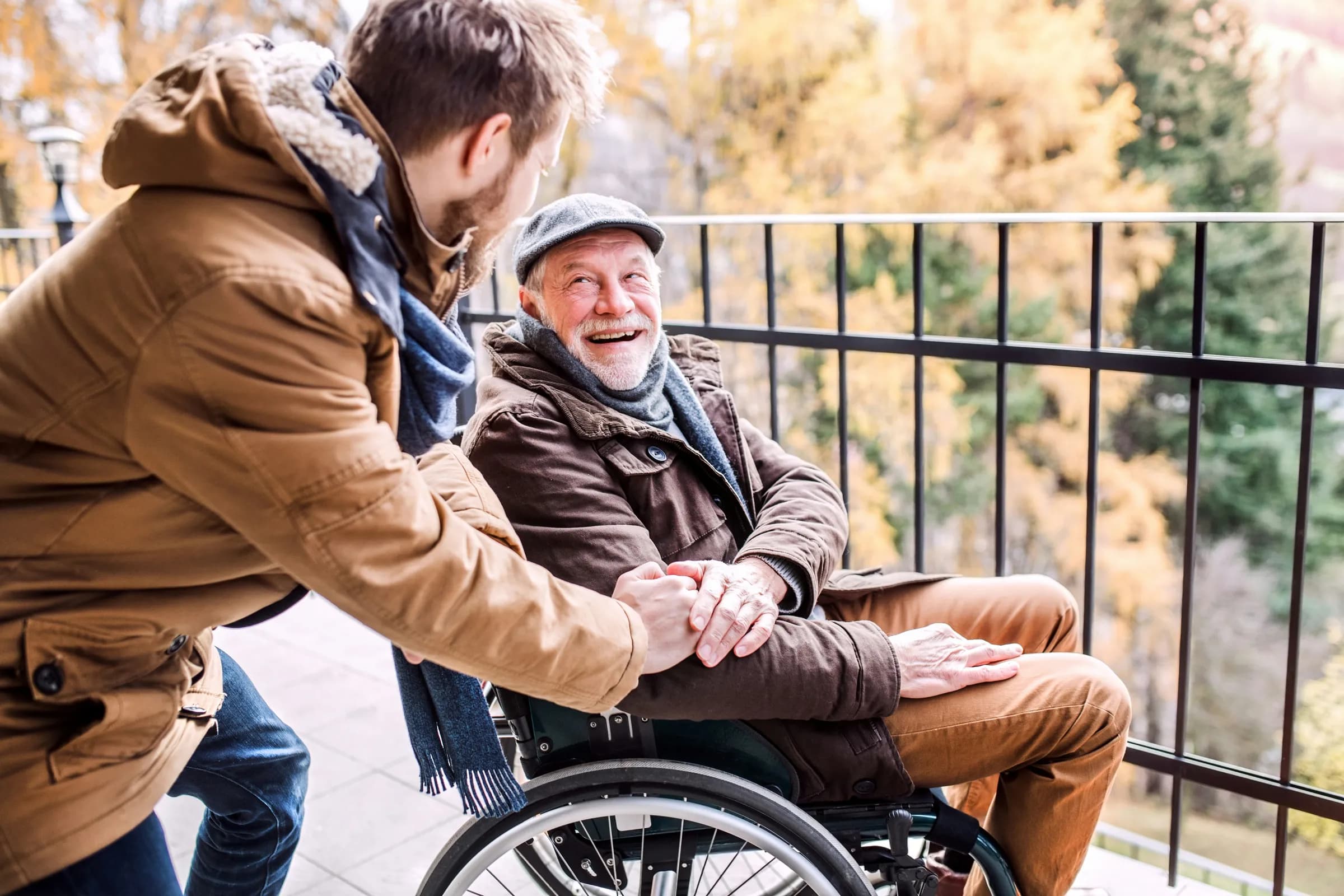 Un homme tient les mains de son père âgé en fauteuil roulant sur une terrasse, un geste de proche aidant